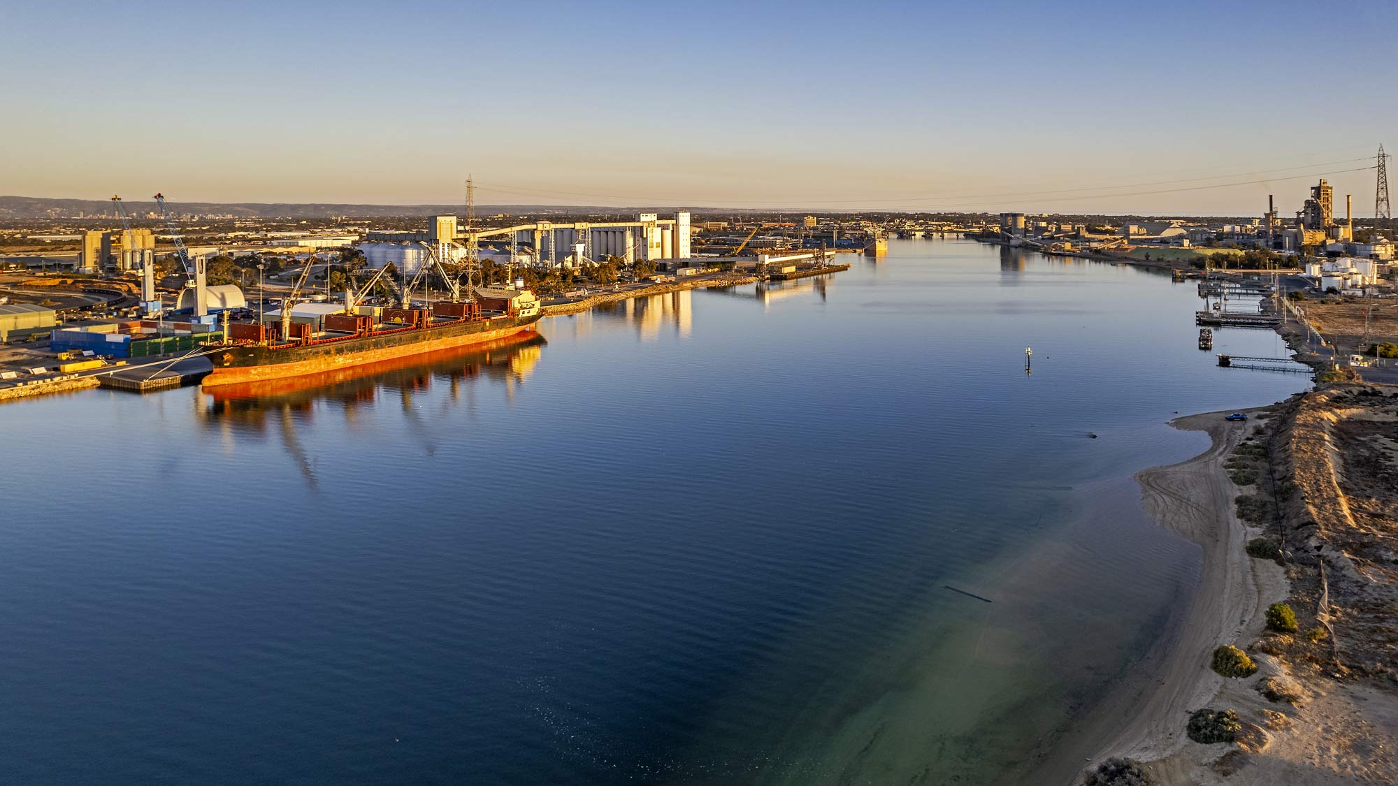 Aerial panoramic view of the Port River docks and industrial district at sunset. Three bulk carriers are visible with the closest loading iron ore. On the right, the cement works and fuel storage tanks and high voltage power lines stretch across the river from huge pylons. In the foreground, the shallow waters, a sandy beach provide a contrast to the dredged shipping lanes.