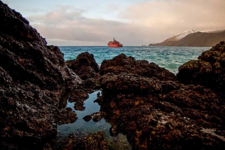 Red boat on horizon of blue Antarctica water with rocky coastline in foreground