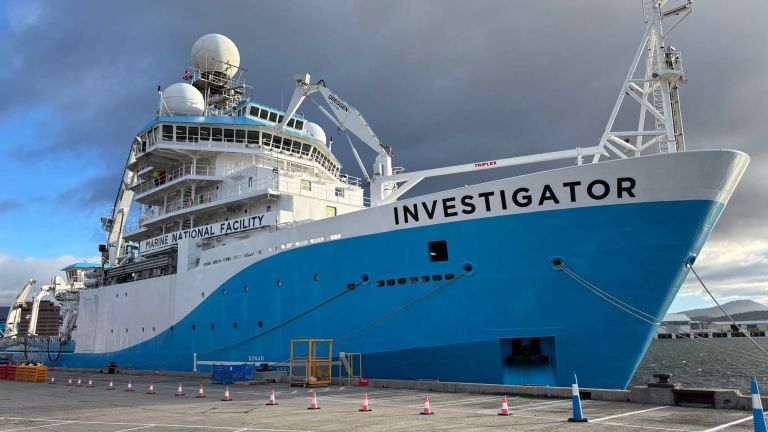 CSIRO Research Vessel Investigator moored at a Hobart dock prior to departure. The large blue-and-white ship fills most of the frame, with scientific equipment visible on the bridge.