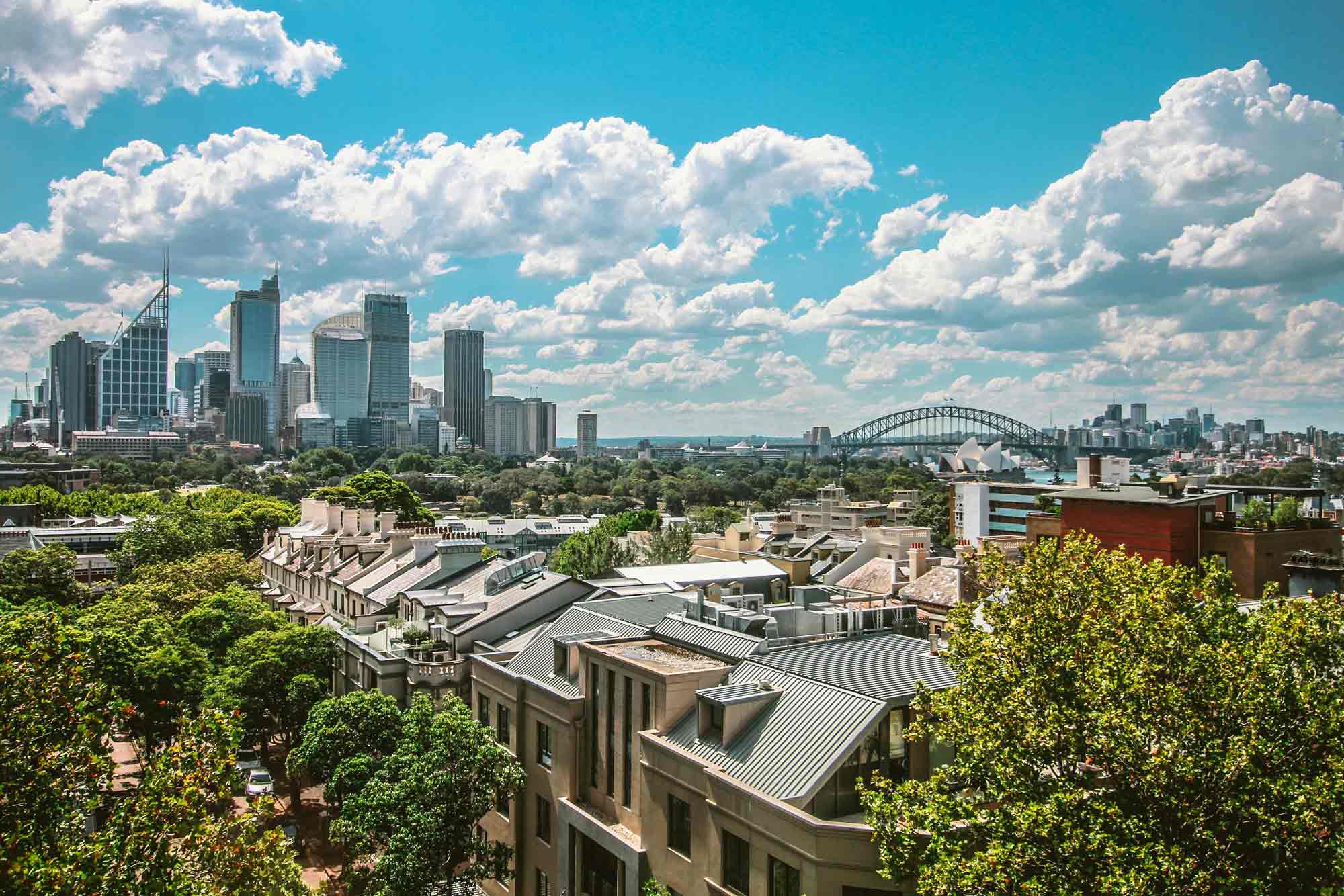 Photograph of Sydney city skyline featuring modern skyscrapers, historic buildings, and lush greenery under a partly cloudy blue sky. Notable landmarks include Sydney Harbour Bridge and Sydney Opera House in the background, highlighting urban development alongside natural elements.