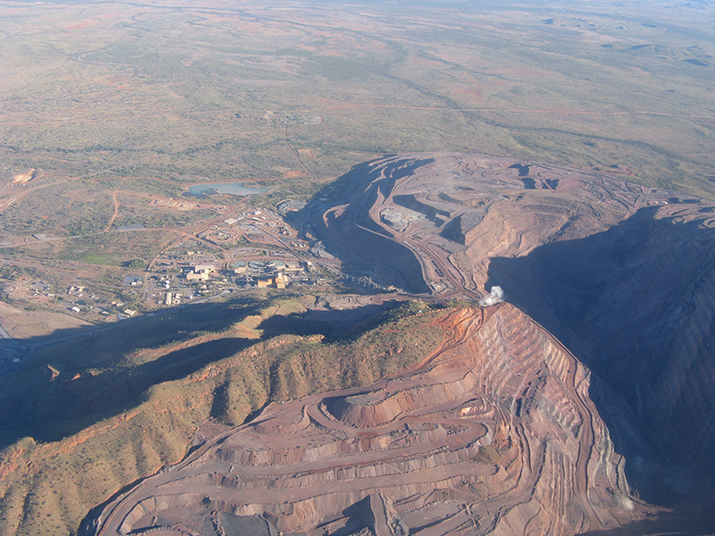  Open pit mine comprised of an excavated hill side with multiple levels and dirt roads. There are buildings behind the hill