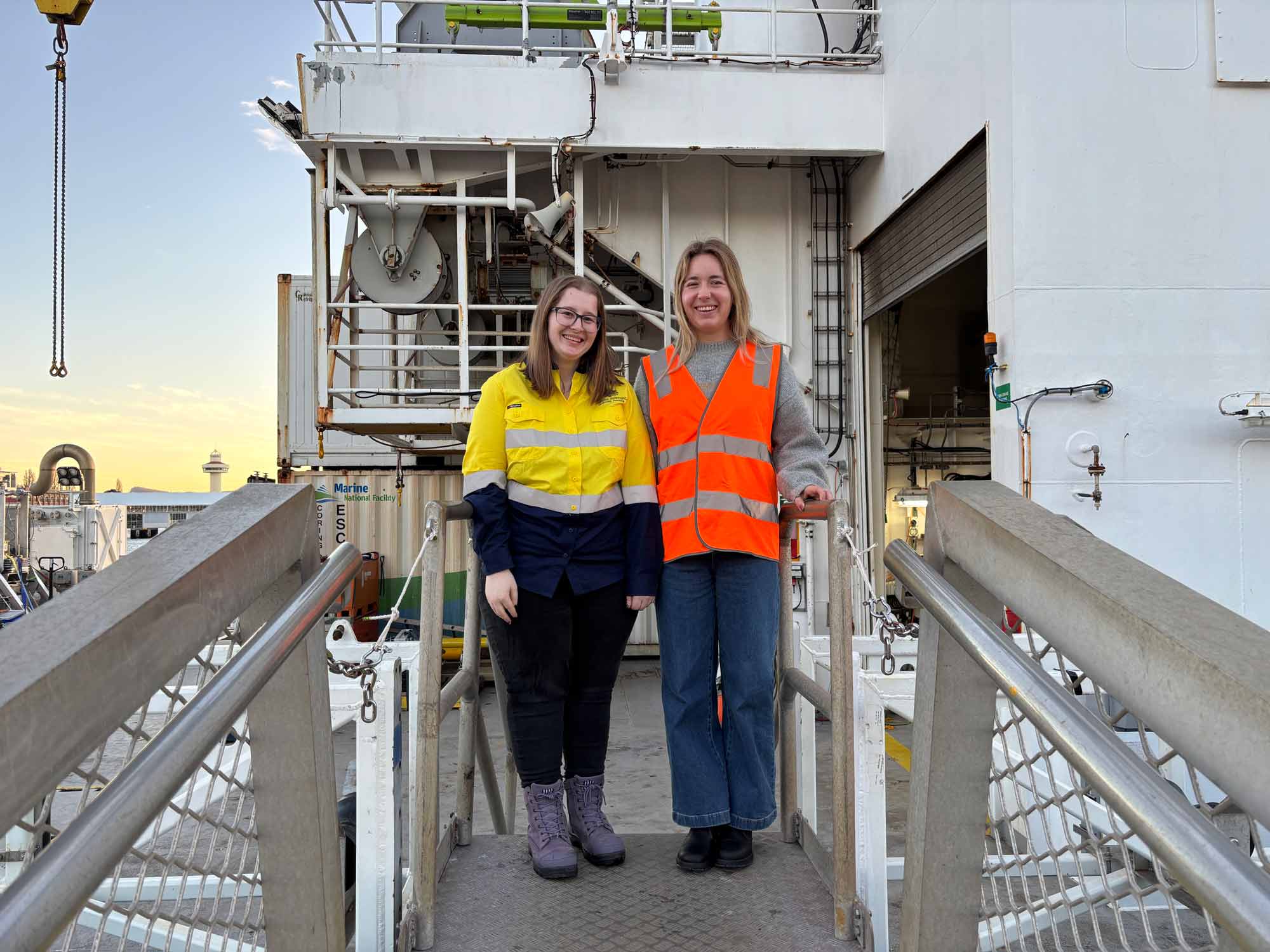 Geoscience Australia Marine Geophysicist Steph Joyce and Marine Geoscientist Kate Purdon aboard the Research Vessel Investigator, wearing high-visibility safety clothing and surrounded by the ship’s deck equipment.