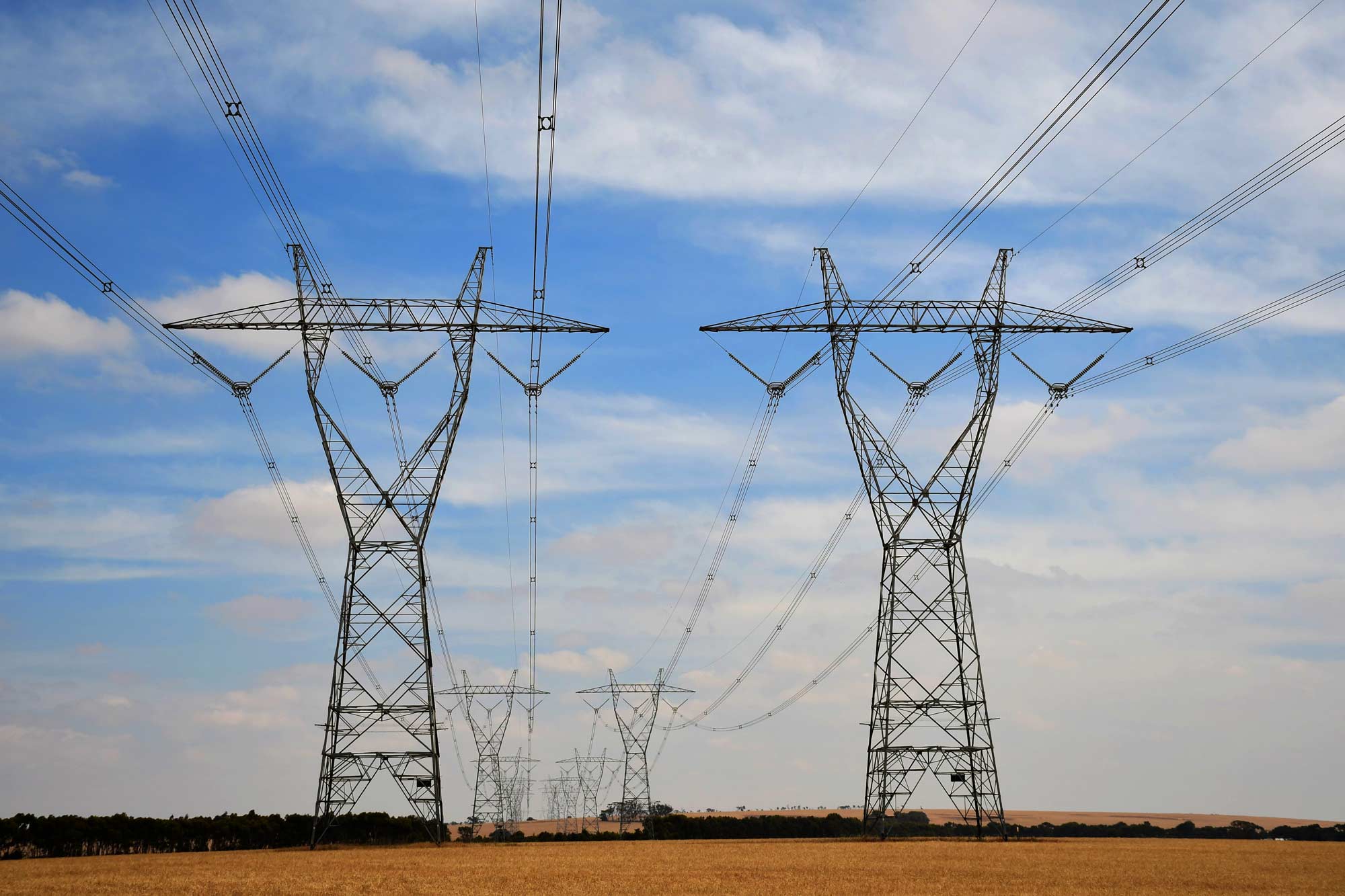 Photograph showing a series of tall electrical transmission towers carrying high-voltage power lines across a flat, open landscape under a partly cloudy sky. The image highlights infrastructure for long-distance electricity distribution with multiple towers aligned in perspective, emphasizing scale and connectivity.