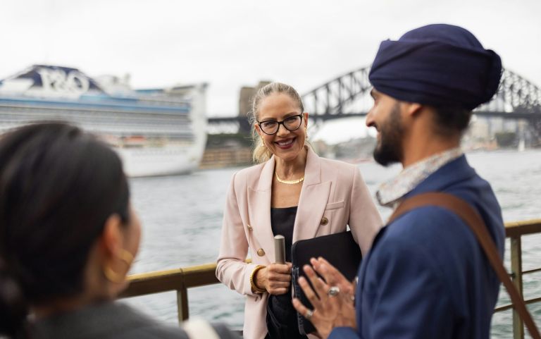 Three people talking on a waterfront promenade in Sydney, with the Sydney Harbour Bridge and a passenger ship visible in the background