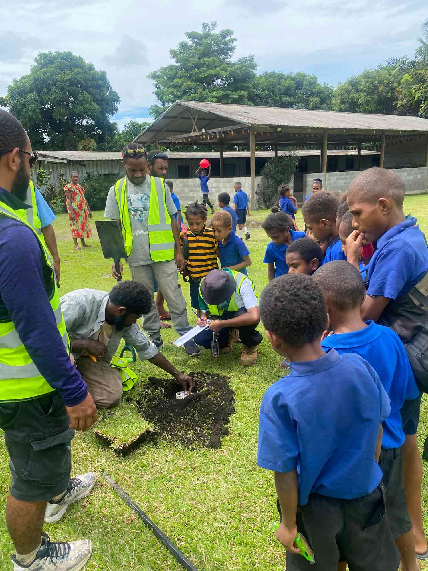 Outdoor scene where four people in high-visibility clothing dig a small hole in the ground, while a group of school children look on. In Lae, Papua New Guinea