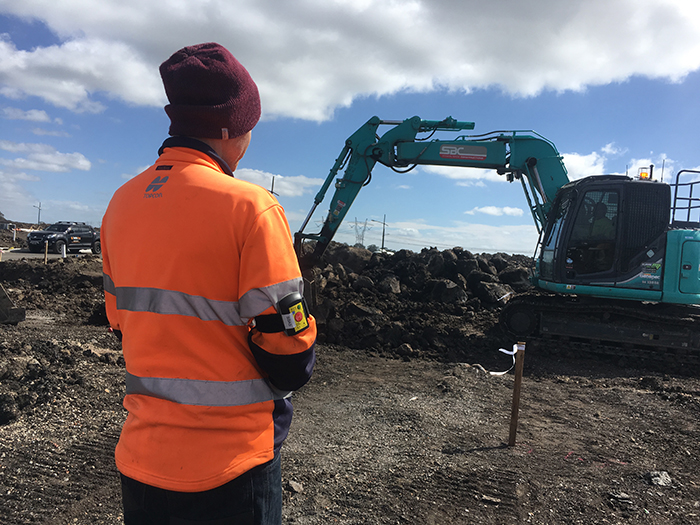 A man is standing approximately 10 metres away from an excavator at a construction site. A small tracking system is strapped his arm.