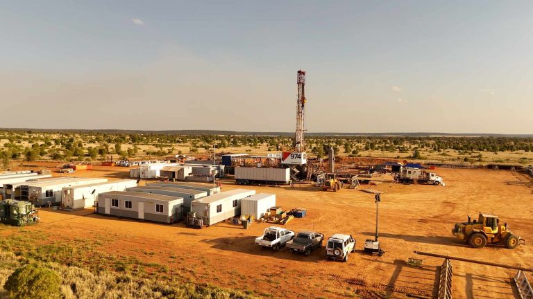 Photograph of a drilling site in a dry, flat landscape with scattered vegetation under a clear sky. The site includes a tall drilling rig, multiple white modular buildings, several trucks, and heavy machinery arranged on reddish-brown soil.