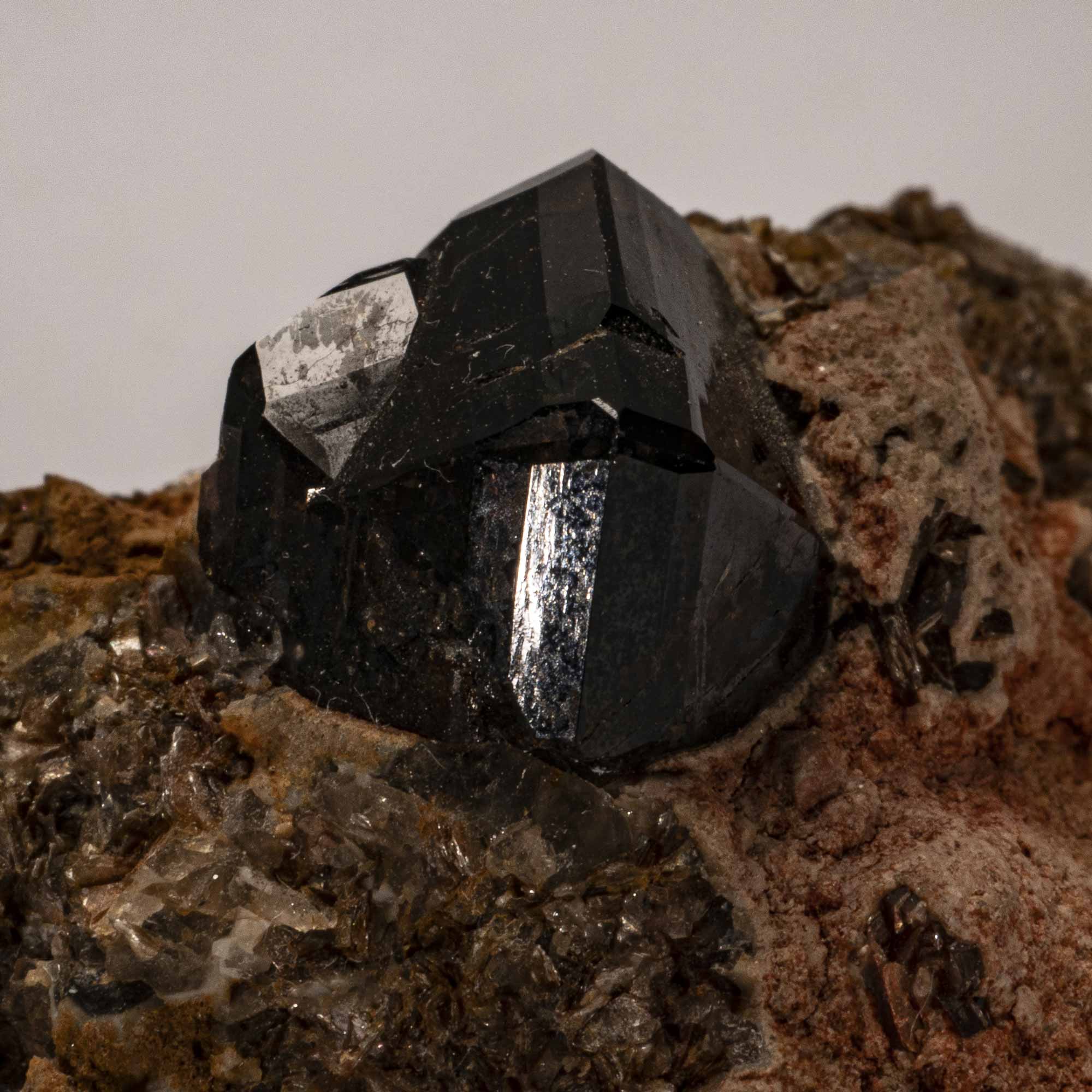 Close-up photograph of a dark, shiny crystal embedded in a rough, brownish rock matrix. The crystal has well-defined geometric faces with reflective surfaces, contrasting with the textured, earthy surrounding material.