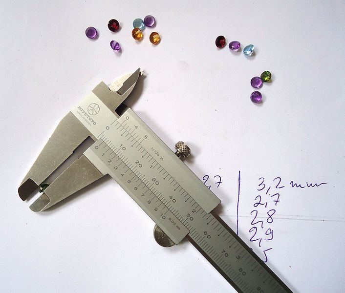 A metal measuring caliper lying on a piece of paper beside coloured, cut rounded gemstones. Some measurements are written on the paper