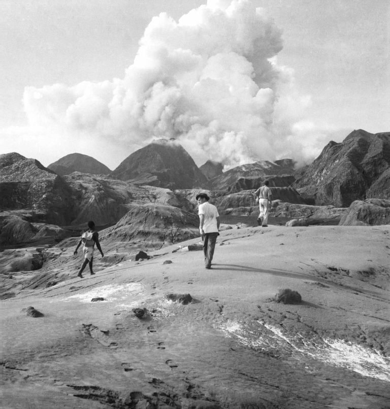 Black and white image of three men walking across a bare rocky landscape walking toward large billowing clouds of smoke that come from a nearby mountain.