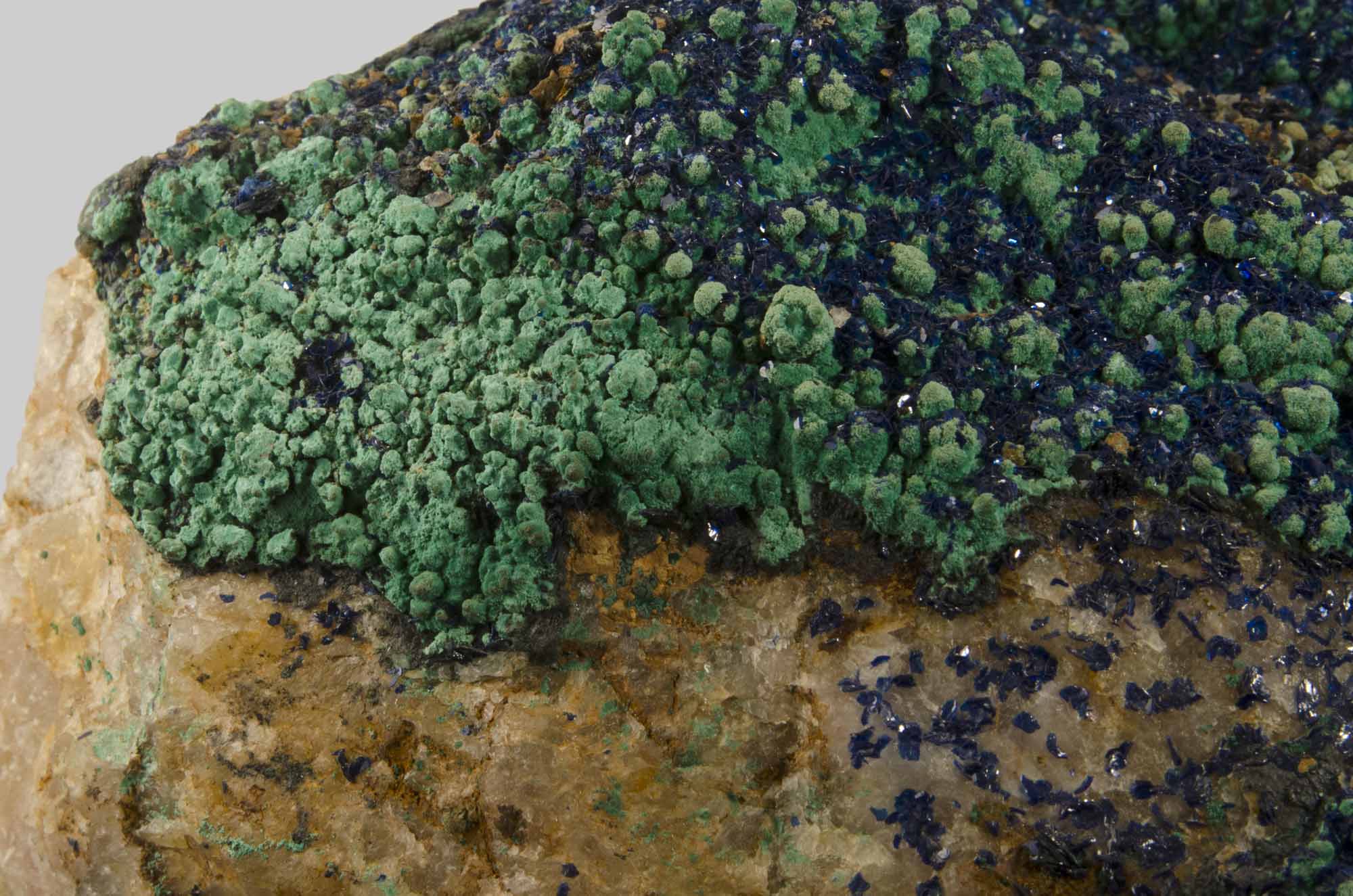 Close-up photograph of a mineral specimen featuring textured green and dark blue crystalline formations on a rough brownish rock surface. The contrast between vibrant green and deep blue crystals highlights mineral diversity and surface irregularities.