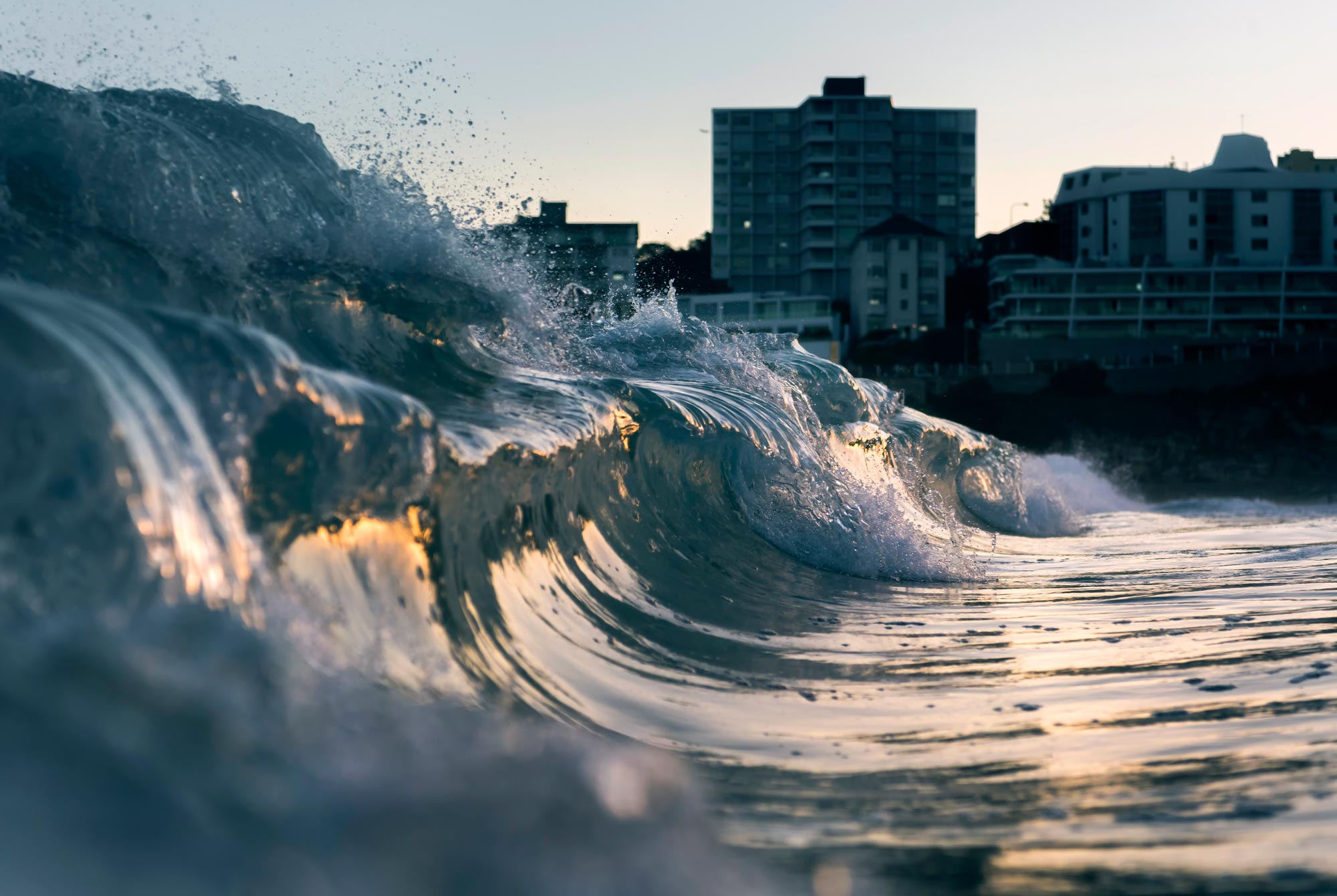 Breaking ocean wave approaching an urban coastline at sunset