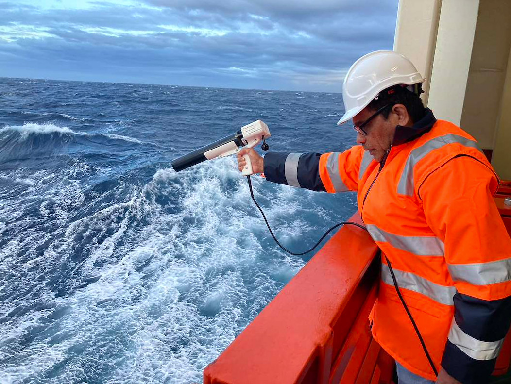 Scientist on the deck of a research vessel using a handheld instrument to measure ocean conditions during a seabed survey at sea