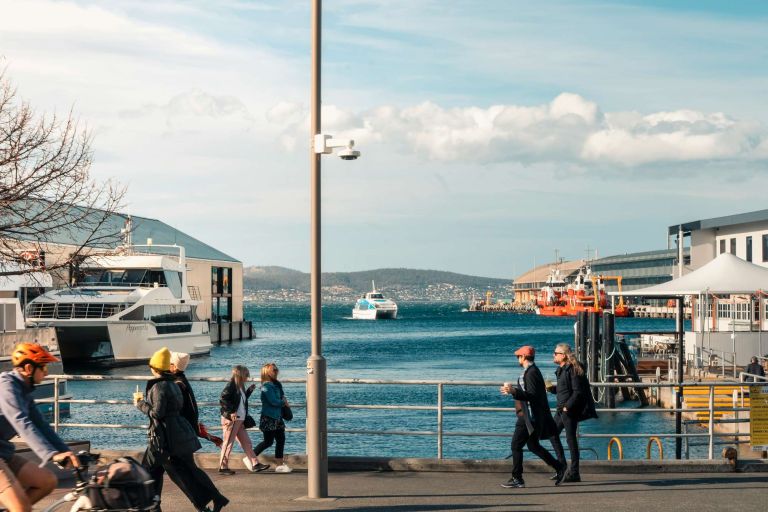 People walking and cycling along a waterfront with ferries docked in the harbour