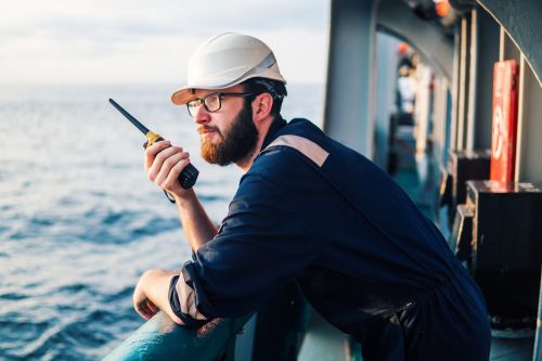 Marine crew member wearing safety equipment uses a handheld radio while working aboard a vessel at sea