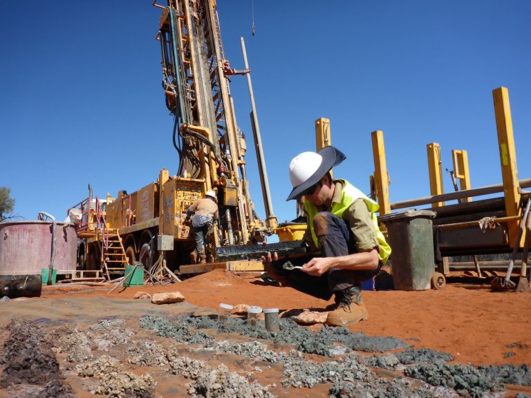 A man in high visibility clothing is crouched down looking at unconsolidated core samples on the ground. Behind the man is another worker at a drill rig