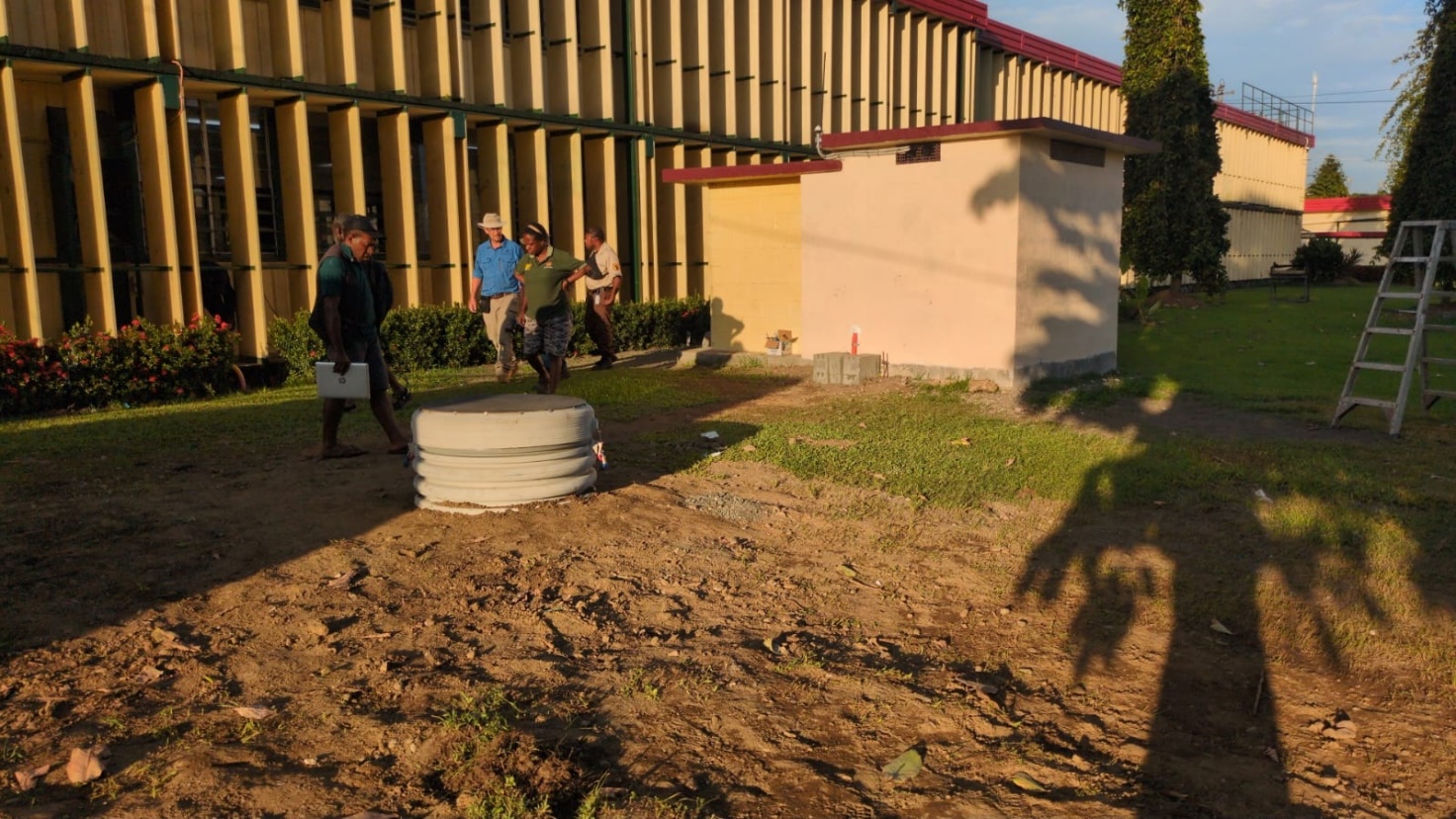 Outdoor scene where four staff inspect a large circular platform that holds earthquake monitoring equipment. Outside the UniTech Lae premises.