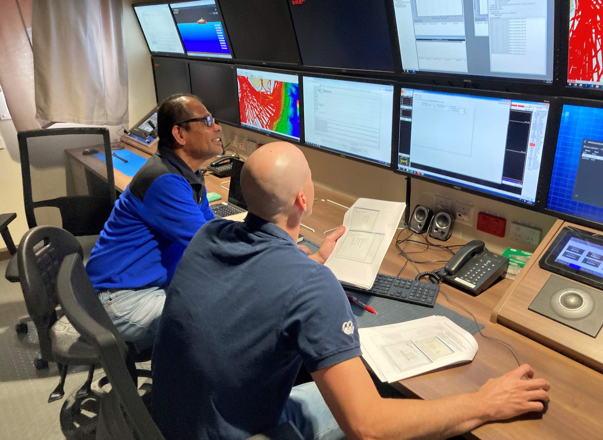 Two scientists monitor and process seabed mapping data on a research vessel, reviewing multibeam survey outputs across multiple computer displays in the ship's operations room