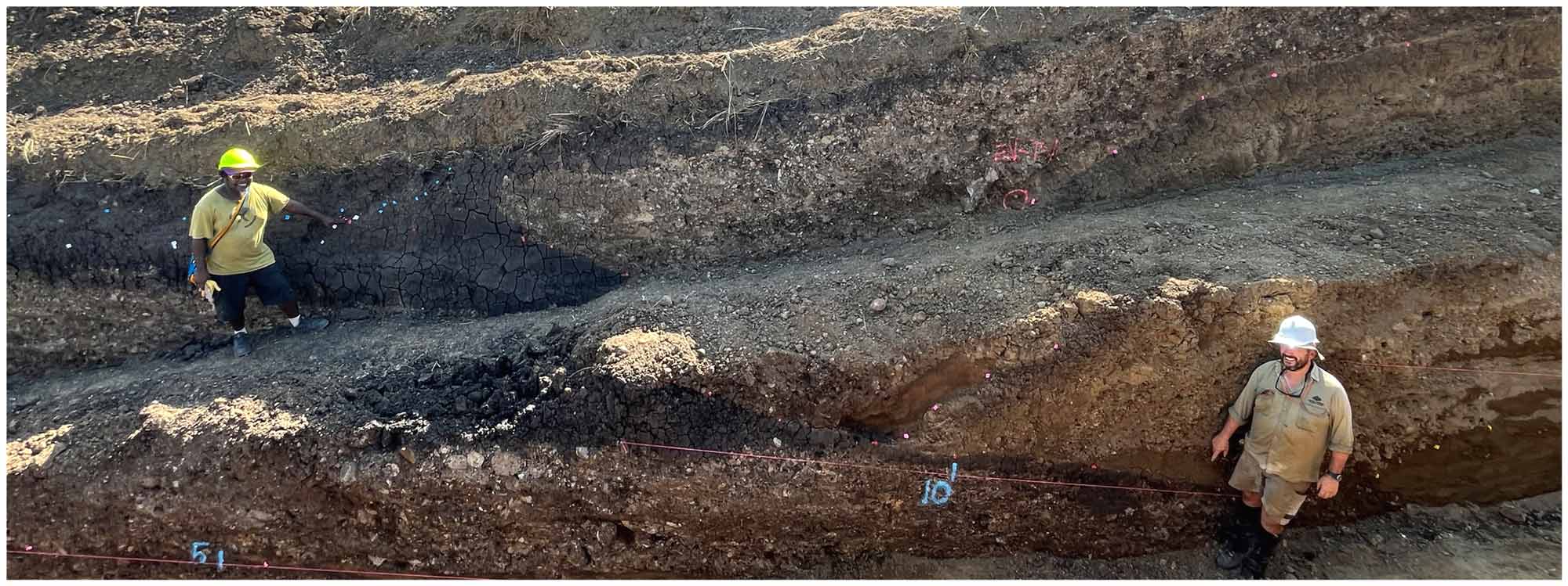 Two people stand in a large trench pointing at different layers in the soil. Near Lae, Papua New Guinea.