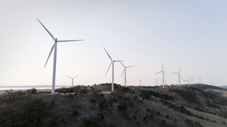 Photograph of a wind farm at dusk featuring multiple wind turbines spread across a dark landscape under a fading sky. The turbines vary in size and are silhouetted against the gradient sky, highlighting renewable energy infrastructure in a natural setting.
