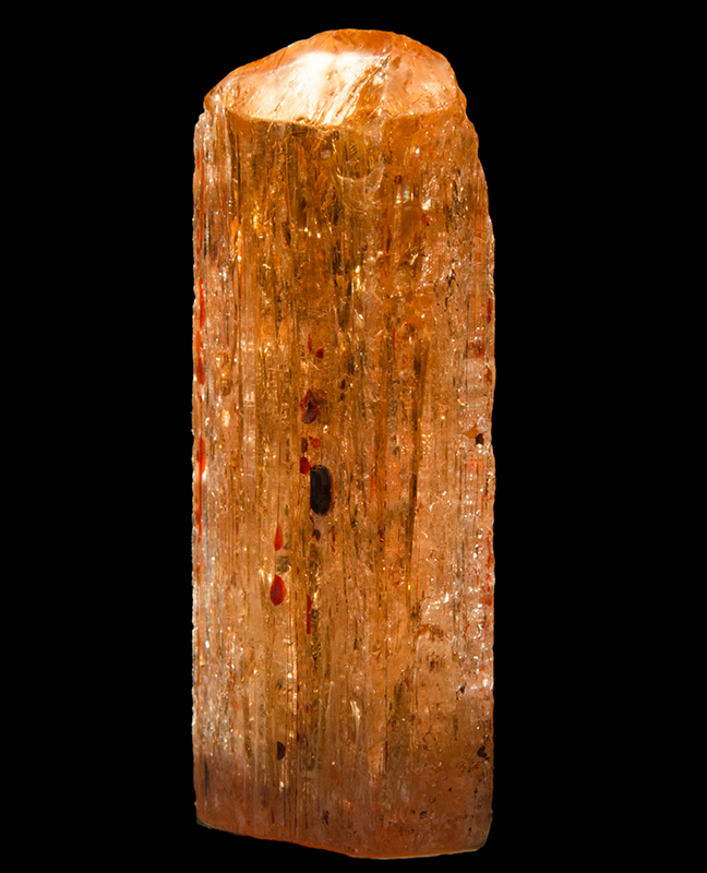 A red coloured topaz crystal with vertical striations and orange and black inclusions in it.