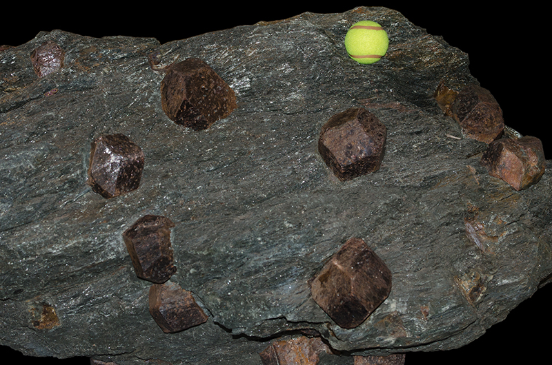 Grey coloured rock with large red crystals in it, sitting on a wooden pallet. The crystals are as large or larger than a tennis ball which has been used as a scale