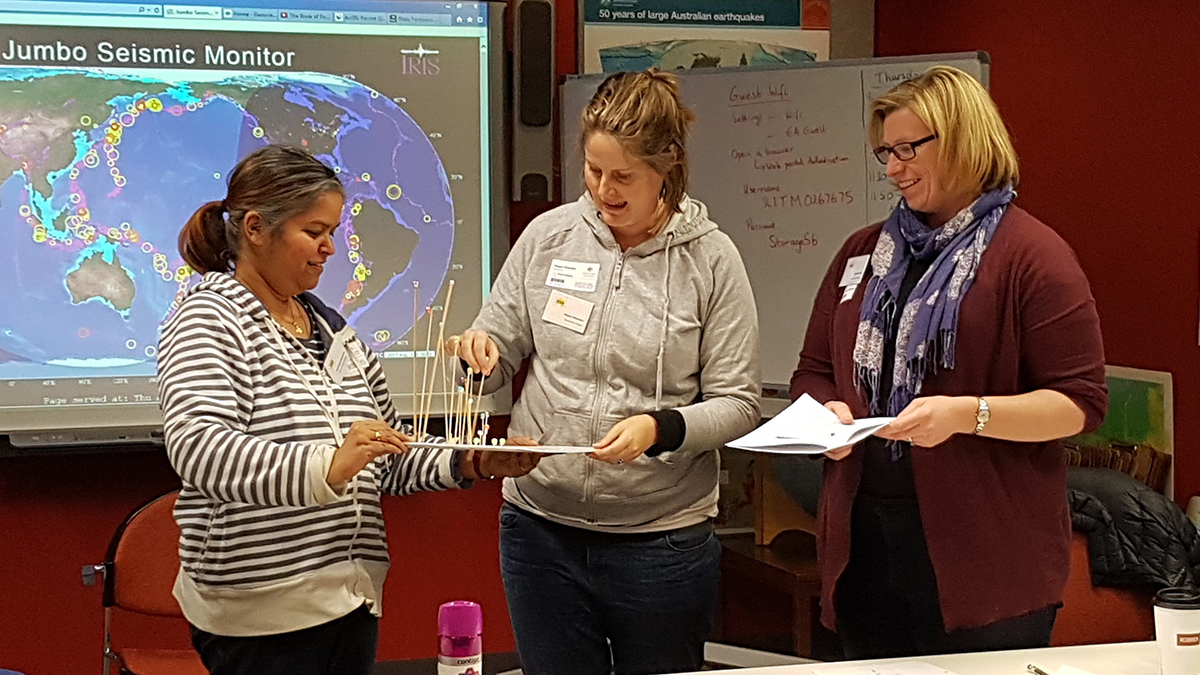 Three teachers explaining a model they have created with a piece of paper, skewers and beads to describe earthquake depth