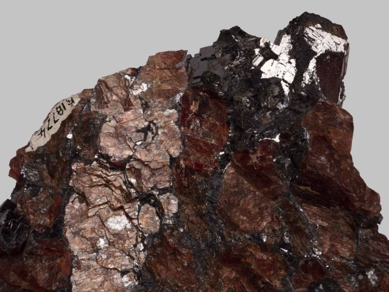 Photograph of a rough mineral specimen featuring metallic, shiny silver and deep reddish-brown crystalline surfaces. The specimen displays a complex texture with reflective patches and a small label attached on the left side.