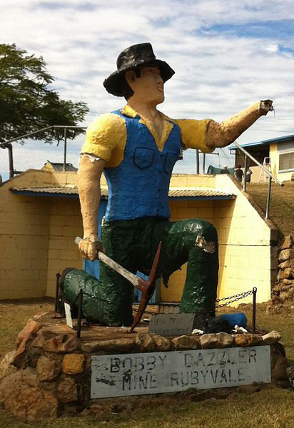Statue of a miner with a pick in front of yellow coloured timber buildings. The label below the statue reads 'Bobby Dazzler Mine Rubyvale'.