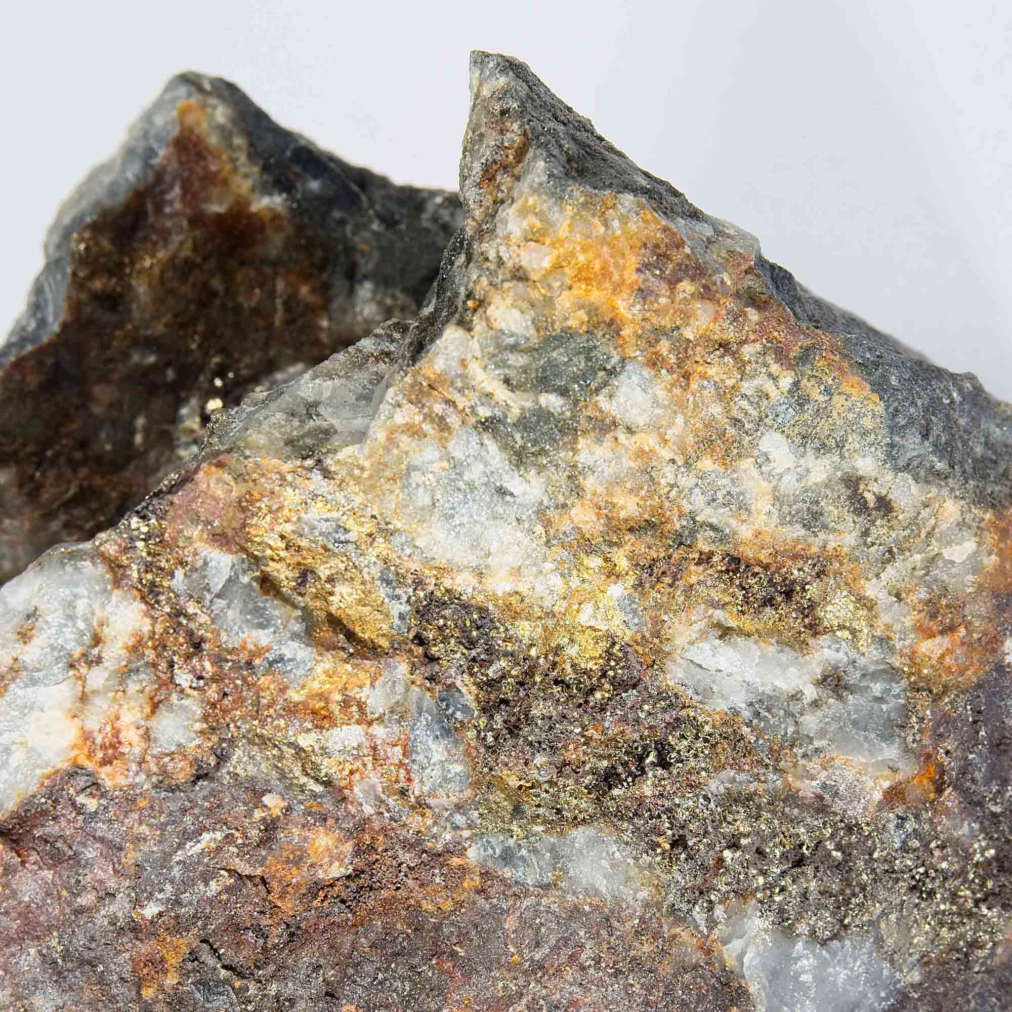 Photograph of a rough mineral rock specimen showcasing a mix of grey, white, yellow, and rusty brown colours with visible crystalline textures. The rock features metallic gold-coloured flecks scattered across its surface, indicating the presence of mineral deposits.