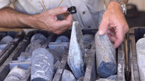 Photograph showing a person examining cylindrical rock core samples arranged in labelled trays, likely for geological or mineral analysis. The person holds a magnifying lens in one hand and points to a specific core sample with the other, highlighting detailed inspection of rock textures and features.
