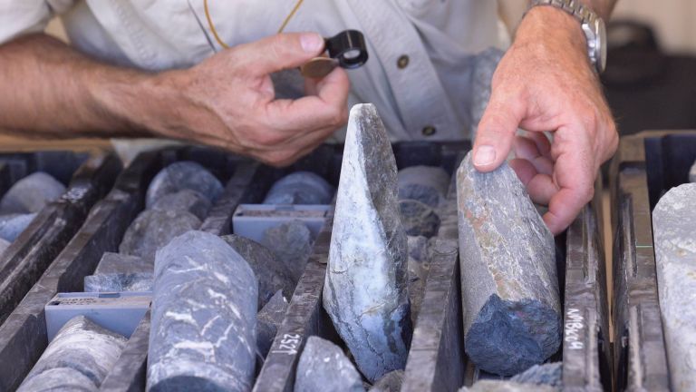 Photograph showing a person examining cylindrical rock core samples arranged in labelled trays, likely for geological or mineral analysis. The person holds a magnifying lens in one hand and points to a specific core sample with the other, highlighting detailed inspection of rock textures and features.