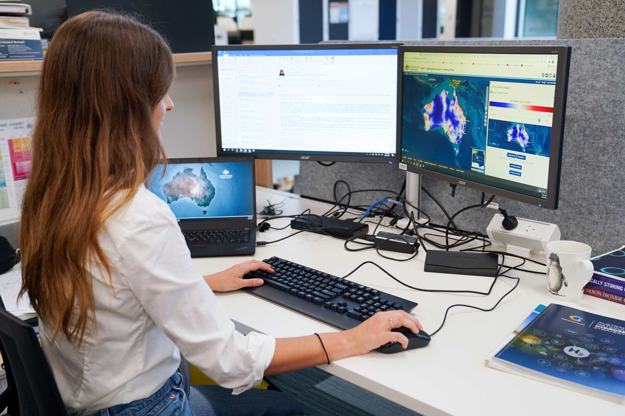 A woman with long dark hair sits at a desk in an office. A computer screen in front of her shows a map of Australia overlaid with scientific data.