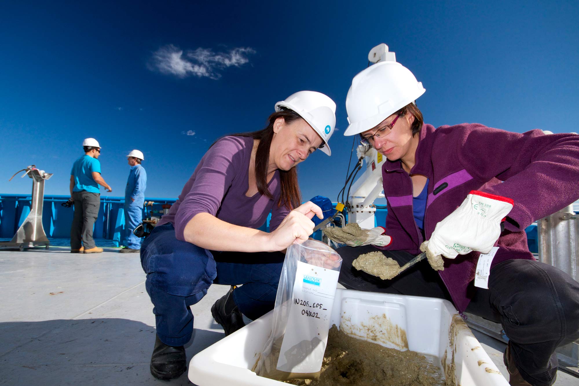 Two women on the deck of a ship wearing hard hats examine mud samples from a white plastic tub.