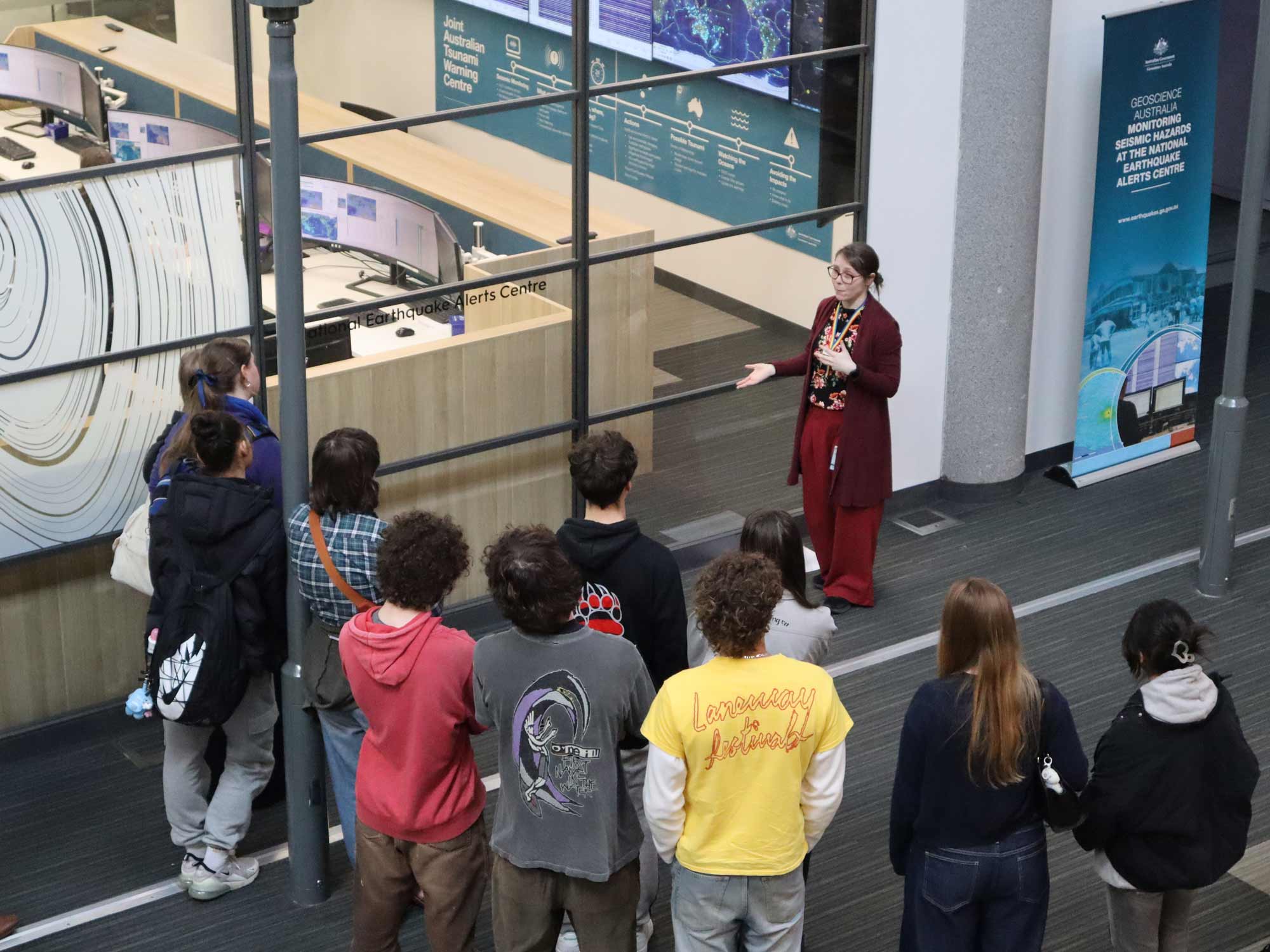 Taken indoors from an upper floor, the image shows a dark haired woman in a long dark red coat standing next to a large window speaking to a group of eleven teenagers, all with their backs to the camera. Through the window can be seen computer displays of maps and scientific data.
