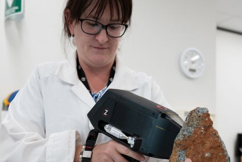 A woman with dark hair and glasses wearing a white lab coat is indoors holding a piece of scientific equipment against a large brown rock.