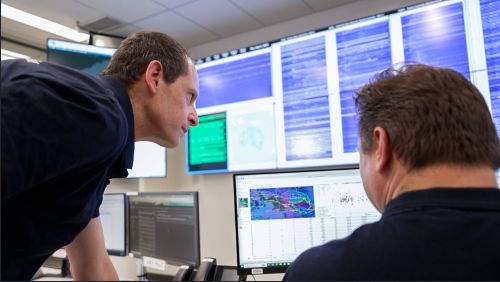 Two men in blue shirts are in front of a monitor showing scientific maps and data. There are screens on the wall in front of them showing multiple wavy lines indicating seismic activity.