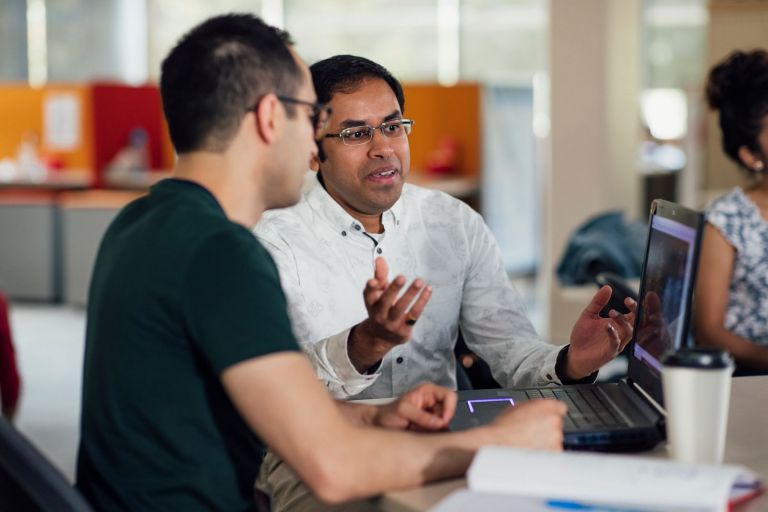 Two men in conversation at a desk with laptop and using hand gestures