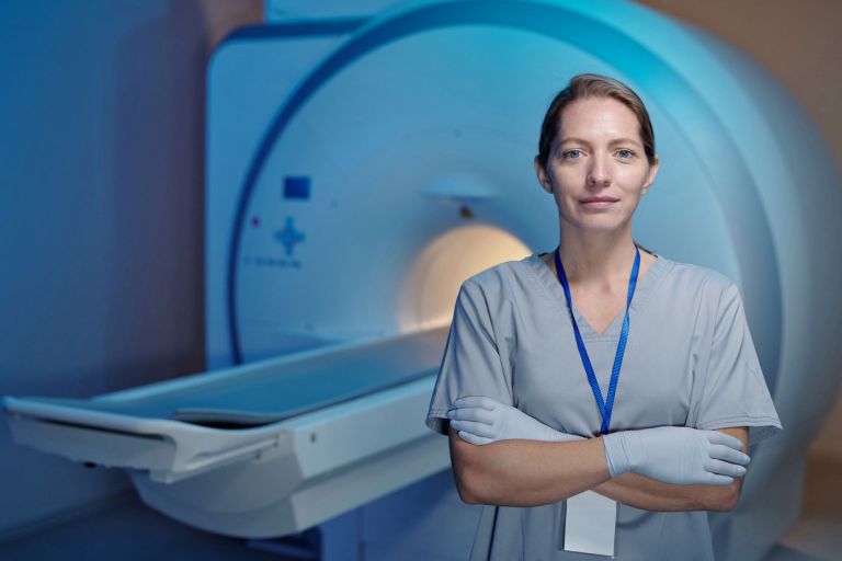 Woman in medical scrubs standing in front of an MRI (magnetic resonance imaging) scanning machine.