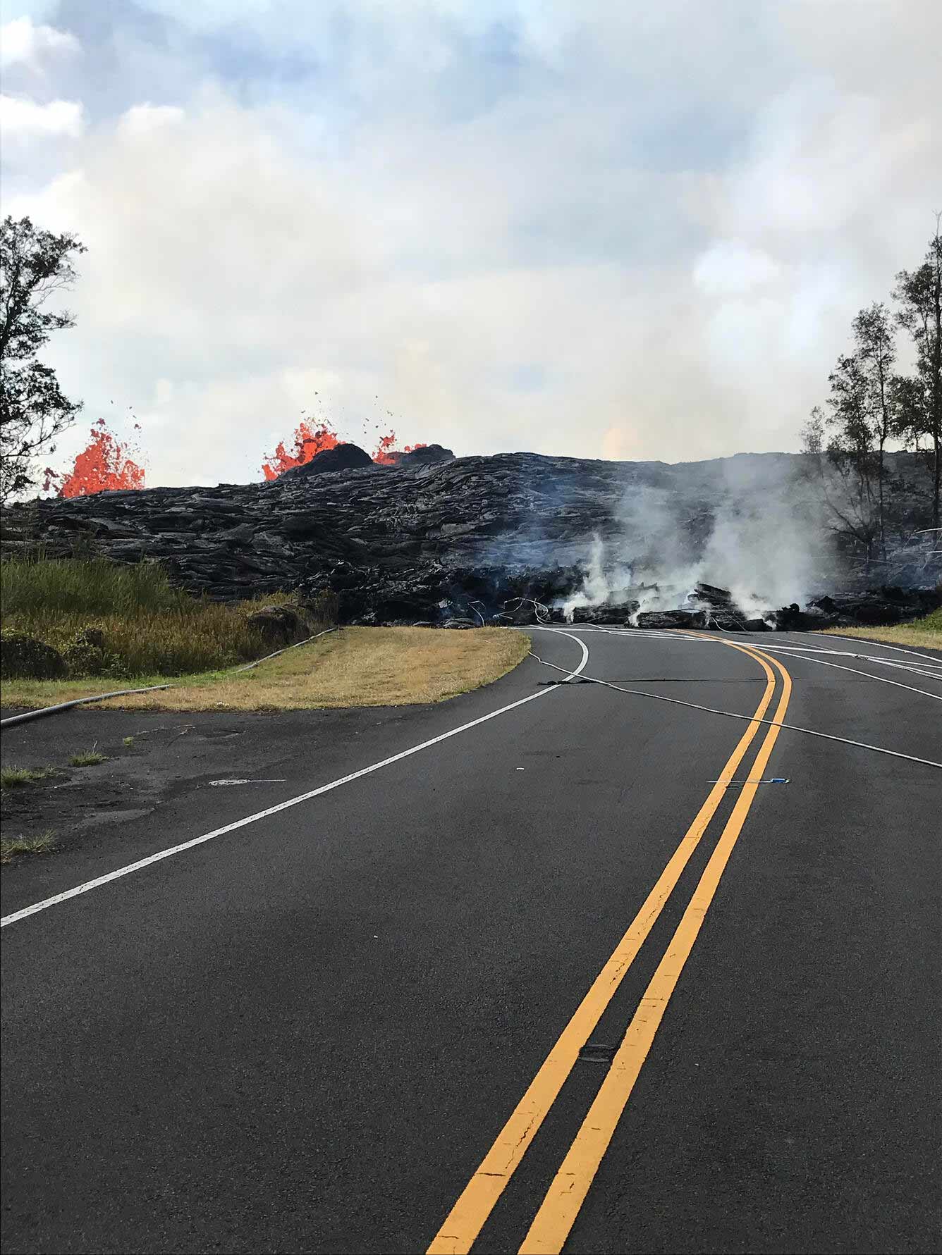 An image showing lava flowing across a road. Molten lava can be seen spurting upwards in the background