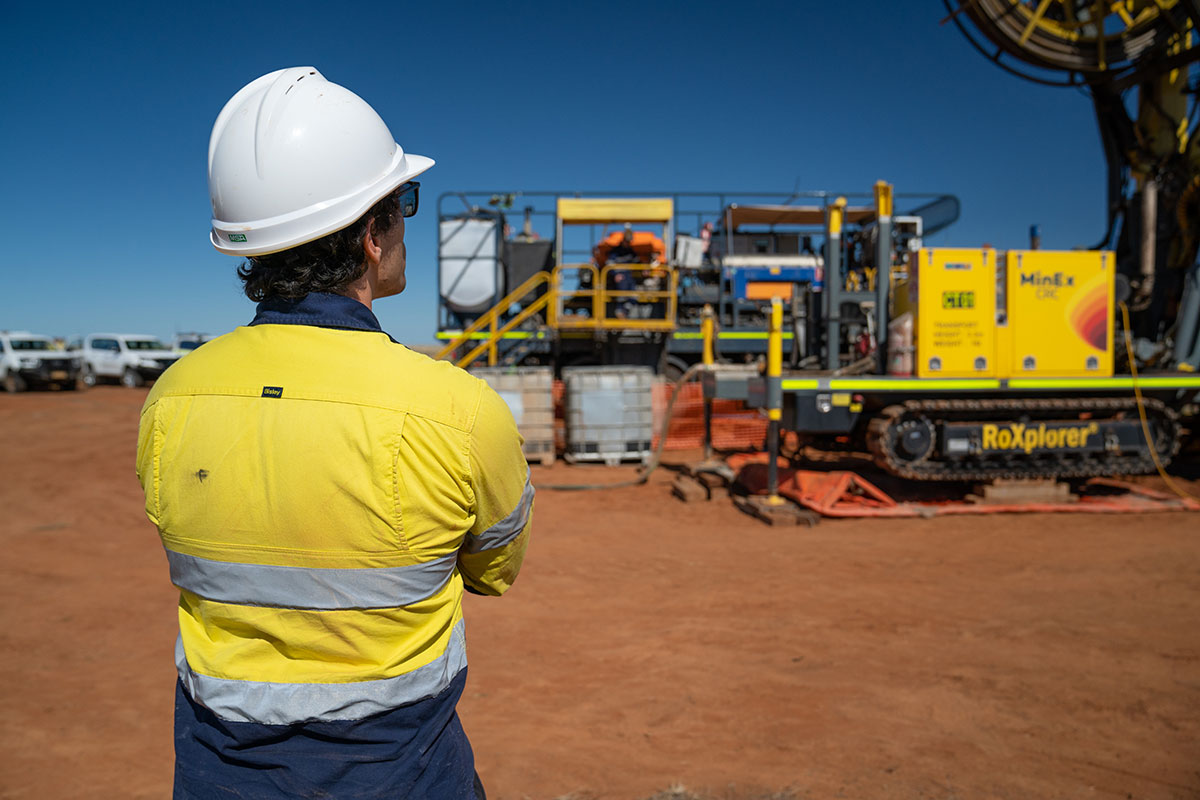 One male geoscientist in a yellow high visibility shirt and white hard hat, with his back to the camera, watches on as the RoXplorer CT drilling platform – a large yellow piece of machinery – is in operation. They are on a site covered in red dirt with a number of white, utility vehicles parked off to left and above is a cloudless blue sky.