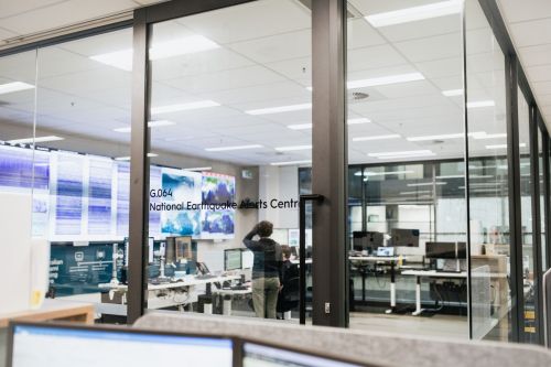 An image from outside the National Earthquake Alert Centre room, that looks through the glass walls at staff standing in front of the wall monitors that display real-time seismic information occurring across Australia.