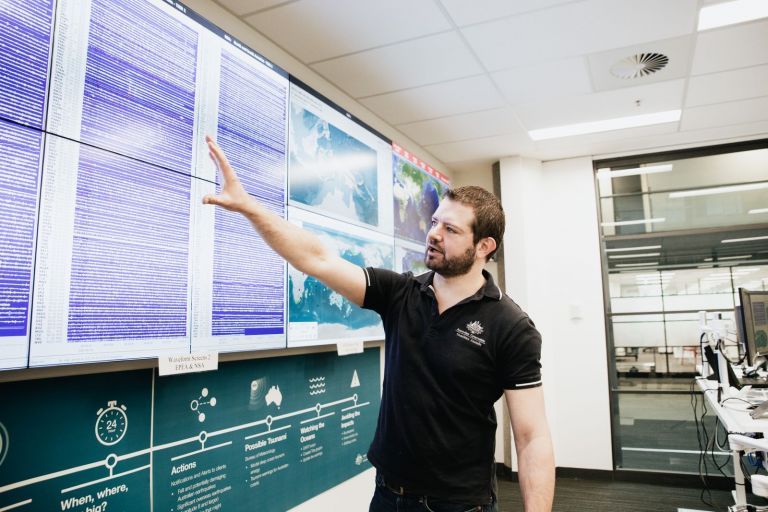 A Geoscience Australia staff member points at the wall monitors in the National Earthquake Alert Centre room, that display real-time seismic information across Australia