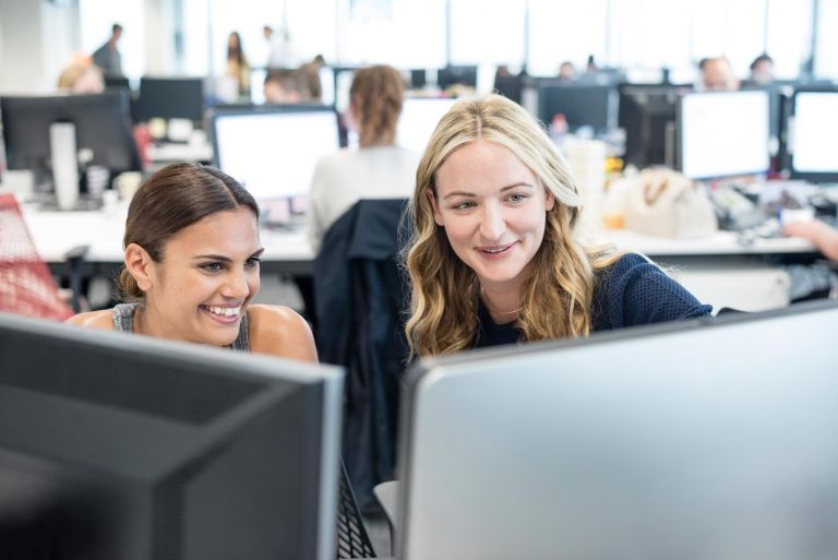 Two young professional women smile at a desktop computer in an open plan office