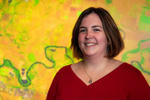 Smiling woman in red top standing in front of yellow satellite image with blue wiggling water