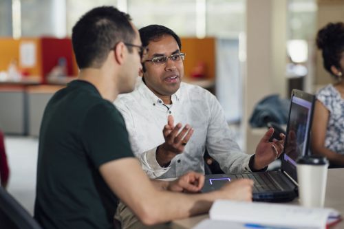 Two men in conversation at a desk with laptop and using hand gestures