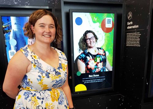 Smiling woman in floral dress stands in front of display that shows her picture titled Earth observation