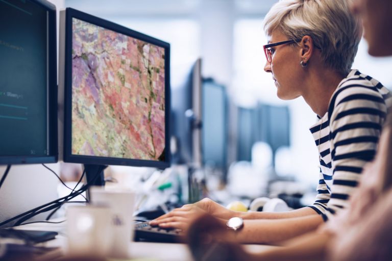 short-haired blonde woman with re glasses looks at code on desktop monitor