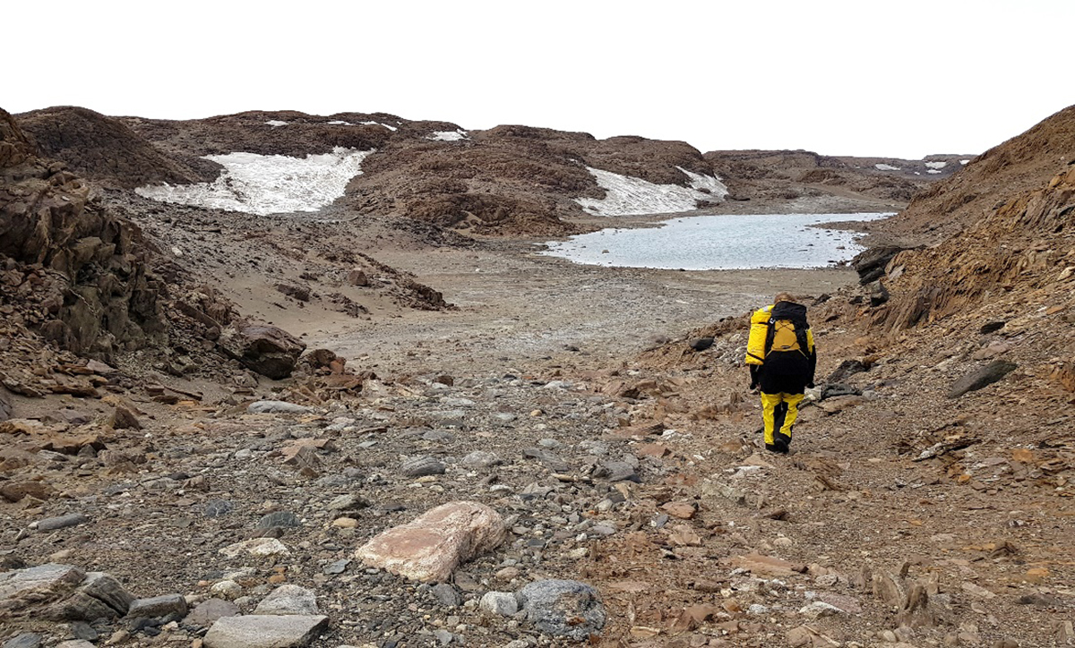 A person with backpack and warm Antarctic clothing walks through a rocky landscape with snow banks and a lake in the background.