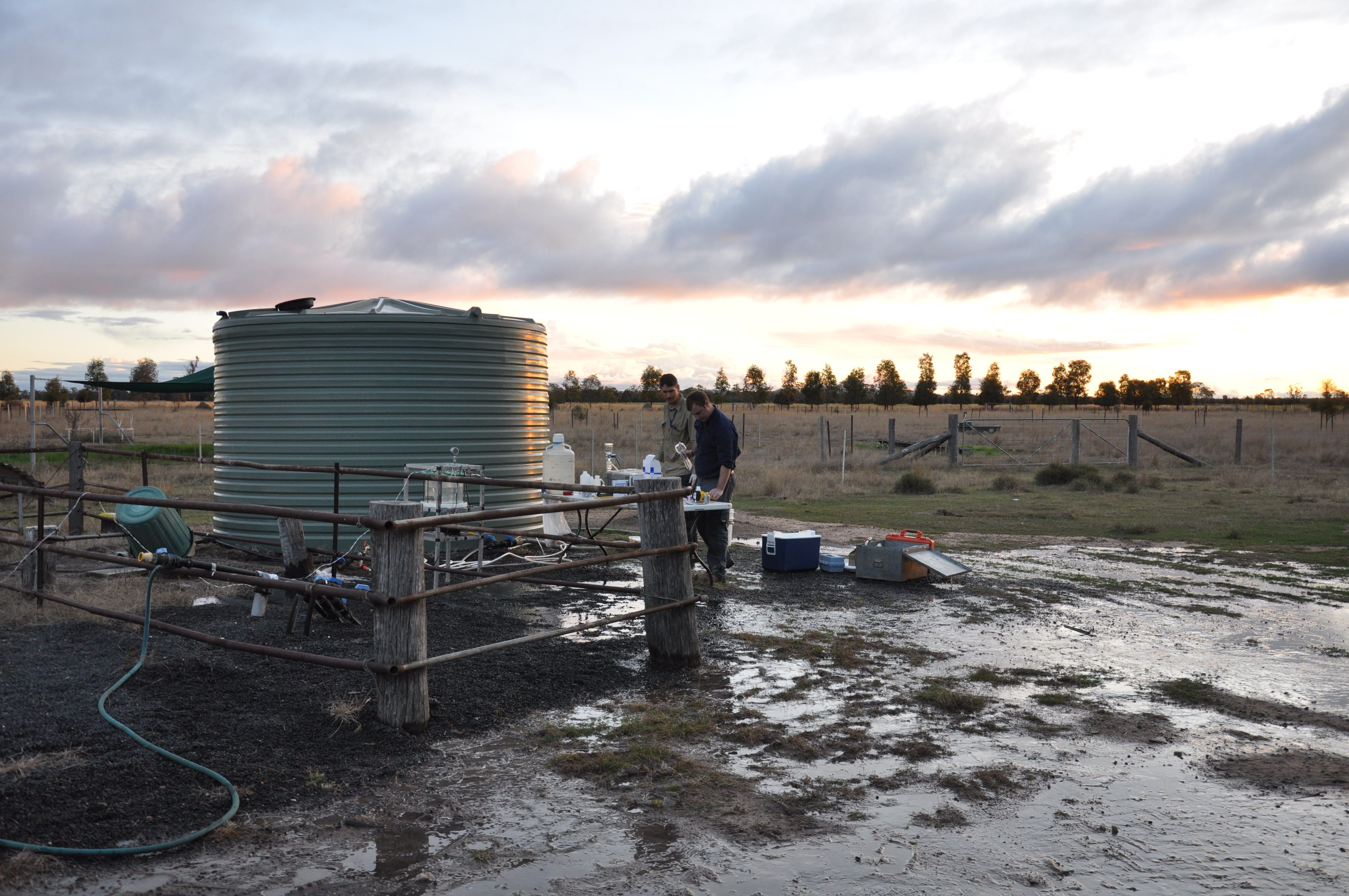 Two men standing near a paddock and a water tank with groundwater gas sampling equipment